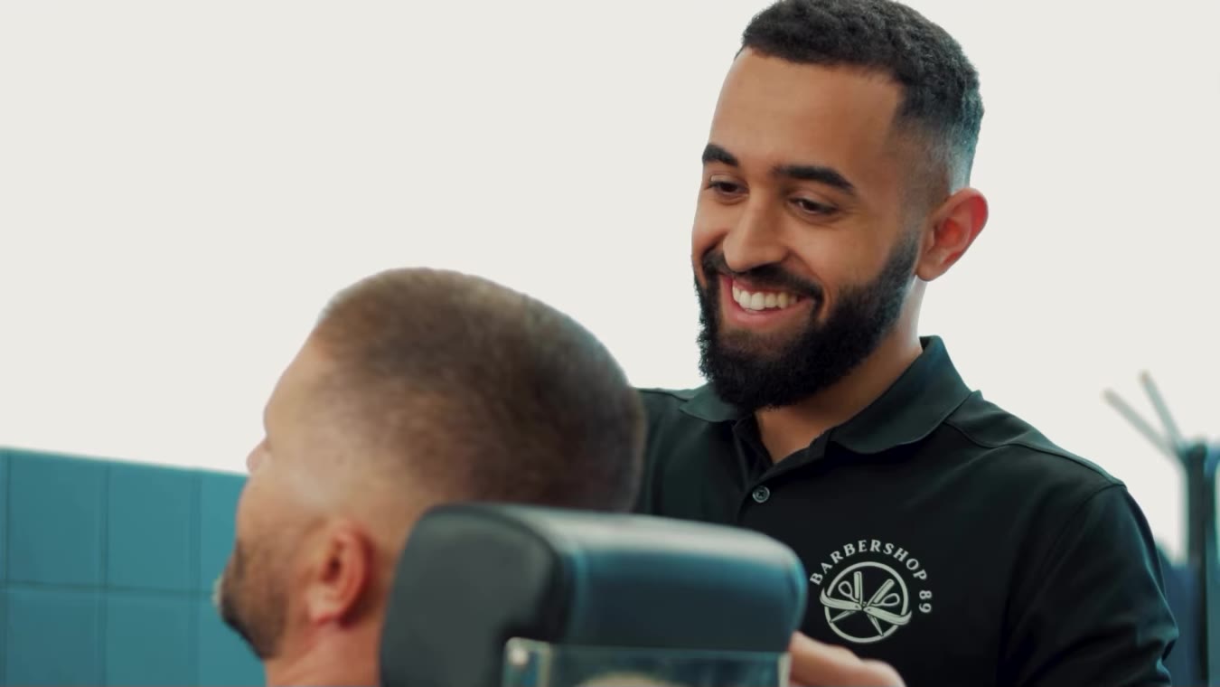 A barber in the black Barbershop 89 polo smiling at a client in the chair